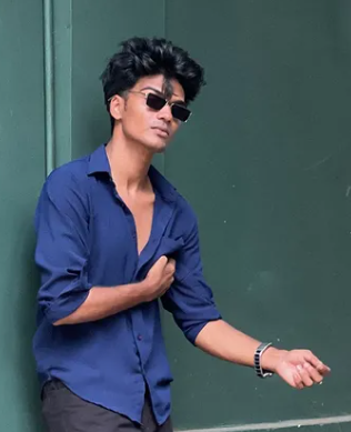 A young man wearing a blue shirt and dark sunglasses, standing casually against a dark green wall, with one arm slightly extended and the other near his chest.
